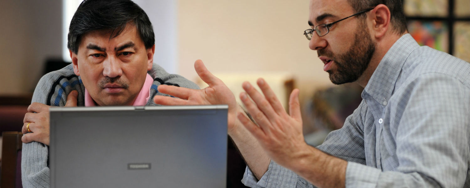 Two men are sitting at a table, facing a laptop computer screen. One man is motioning with his hands as if to explain something.
