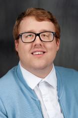 Cody Benedict Headshot. Short, reddish hair with glasses and a light blue cardigan over a white button-up.